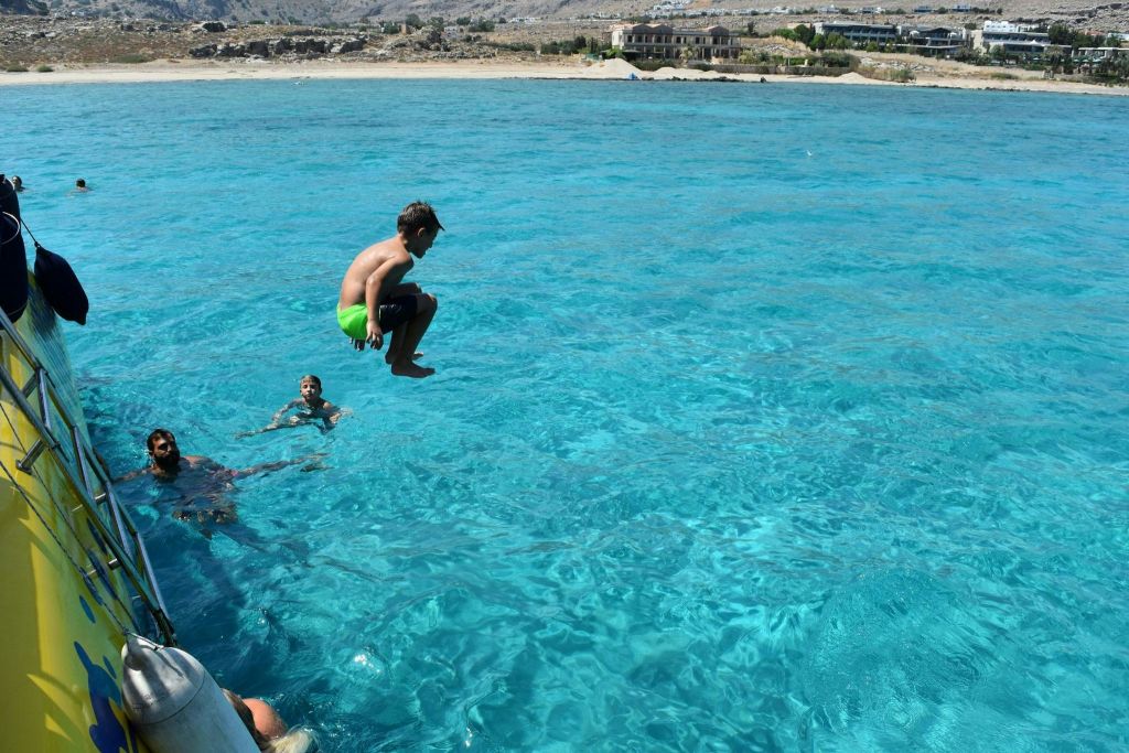 녹색 수영복을 입은 한 소년이 배에서 맑고 푸른 물로 뛰어들고, 다른 소년이 근처에서 수영합니다. 백그라운드에서 해안선을 볼 수 있습니다.