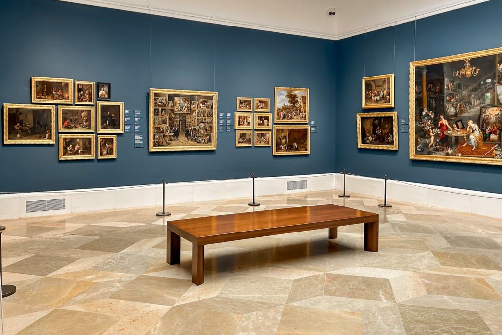 파란색 벽에 금색 액자가 그려진 박물관 갤러리와 중앙의 타일 바닥에 나무 벤치가 있습니다.