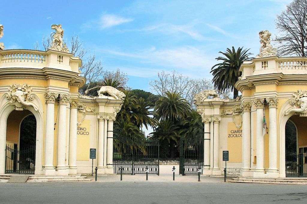 양쪽에 "Giardino Zoologico" 라고 쓰여진 화려한 출입문, 푸른 하늘 아래 조각상과 야자수가 나란히 있습니다.