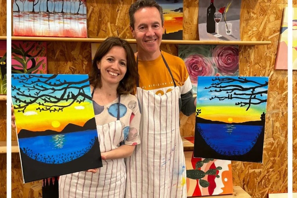 다양한 예술 작품을 배경으로 물 위로 지는 석양의 다채로운 그림을 들고 있는 앞치마를 두 명의 미소 짓는 사람.