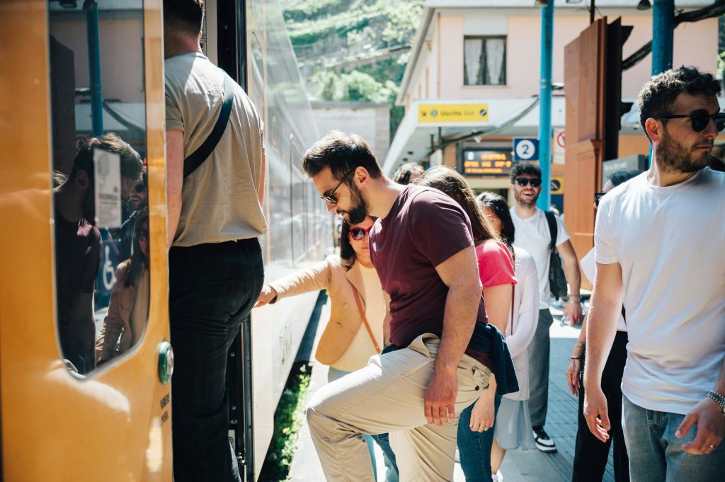 화창한 날 역에서 기차를 타는 사람들. 적갈색 셔츠를 입은 남성과 흰색 셔츠를 입은 남성이 눈에 띄게 보입니다.