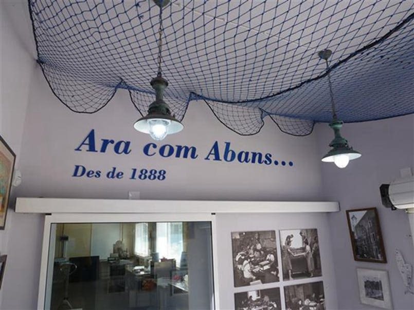 천장에 파란색 그물망, 걸이식 램프, 벽에 "Ara com Abans... Des de 1888" 문자가 있는 실내 객실. 사진은 아래에 표시됩니다.