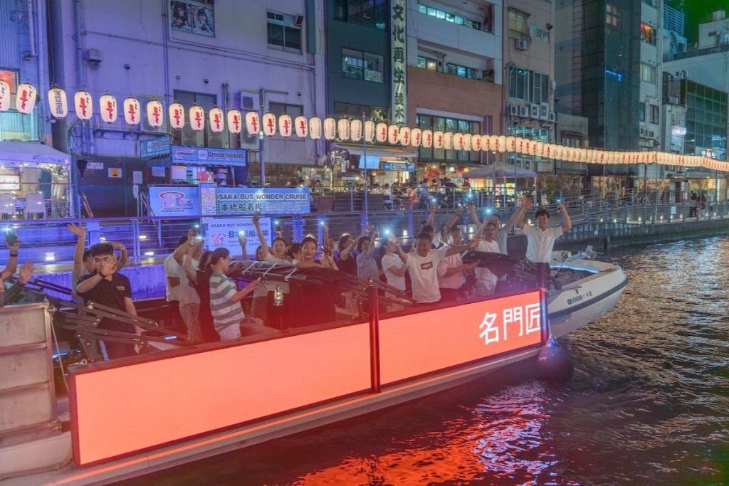 한 무리의 사람들이 밤에 배를 타고 불이 켜진 휴대폰을 들고 등불과 건물을 배경으로 불을 밝힙니다.