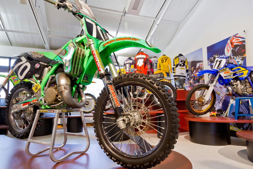 쇼룸에 전시된 초록색 더트 바이크를 클로즈업하여 모토크로스 유니폼과 다른 바이크를 배경으로 합니다.