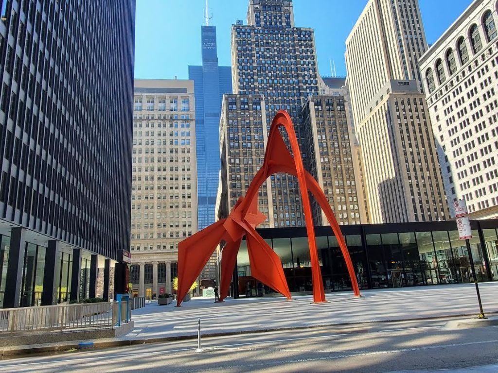 맑고 푸른 하늘 아래 높은 건물로 둘러싸인 광장에 빨간색 추상 조형물이 있습니다.