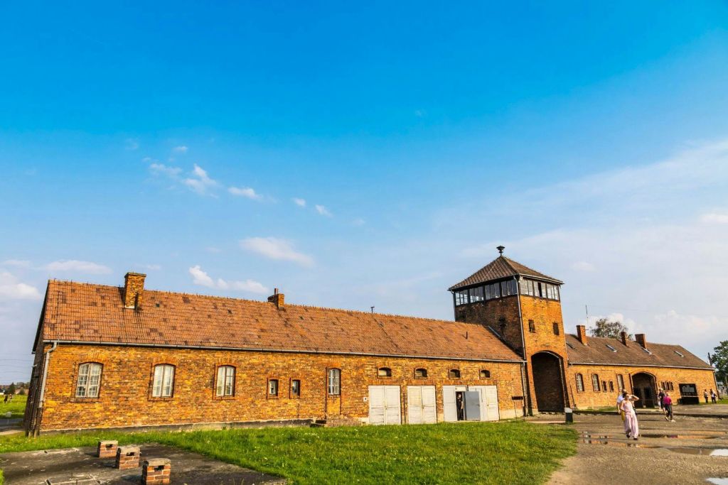 잔디와 포장된 길로 둘러싸인 맑고 푸른 하늘 아래 망루가 있는 벽돌 건물. 일부 문과 작은 창문이 보입니다.