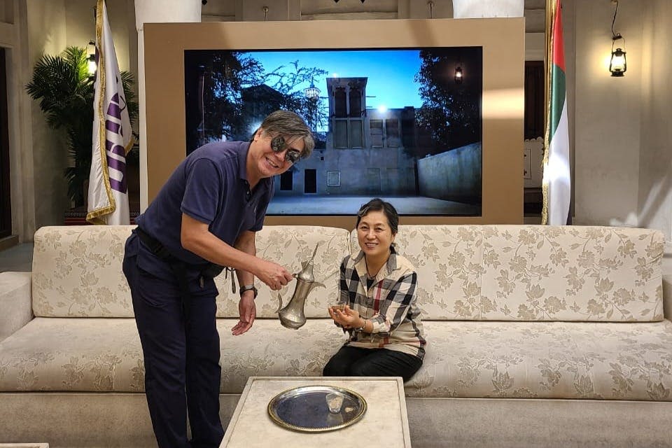 한 남성이 전통적인 커피 포트에서 쏟아져 나오고 한 여성이 소파에 앉아 컵을 들고 있습니다. 아랍에미리트 국기와 TV가 배경입니다.