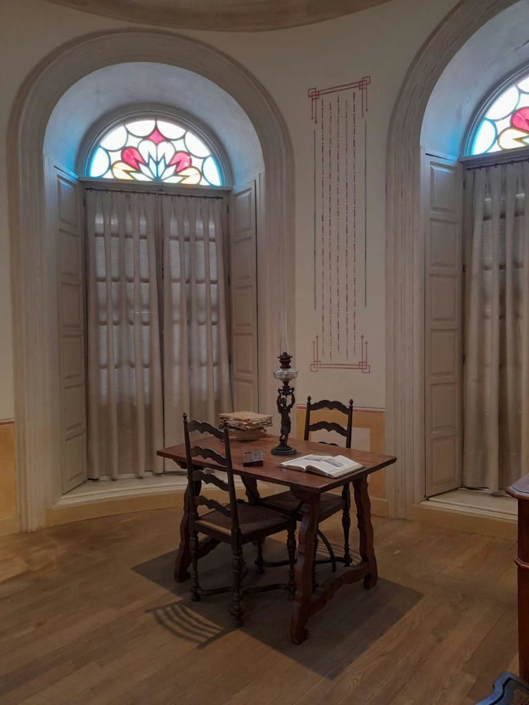 의자 2개, 촛대, 책, 열린 책이 놓인 작은 나무 테이블이 스테인드글라스로 장식된 커튼이 있는 두 개의 창문 사이에 놓여 있습니다.