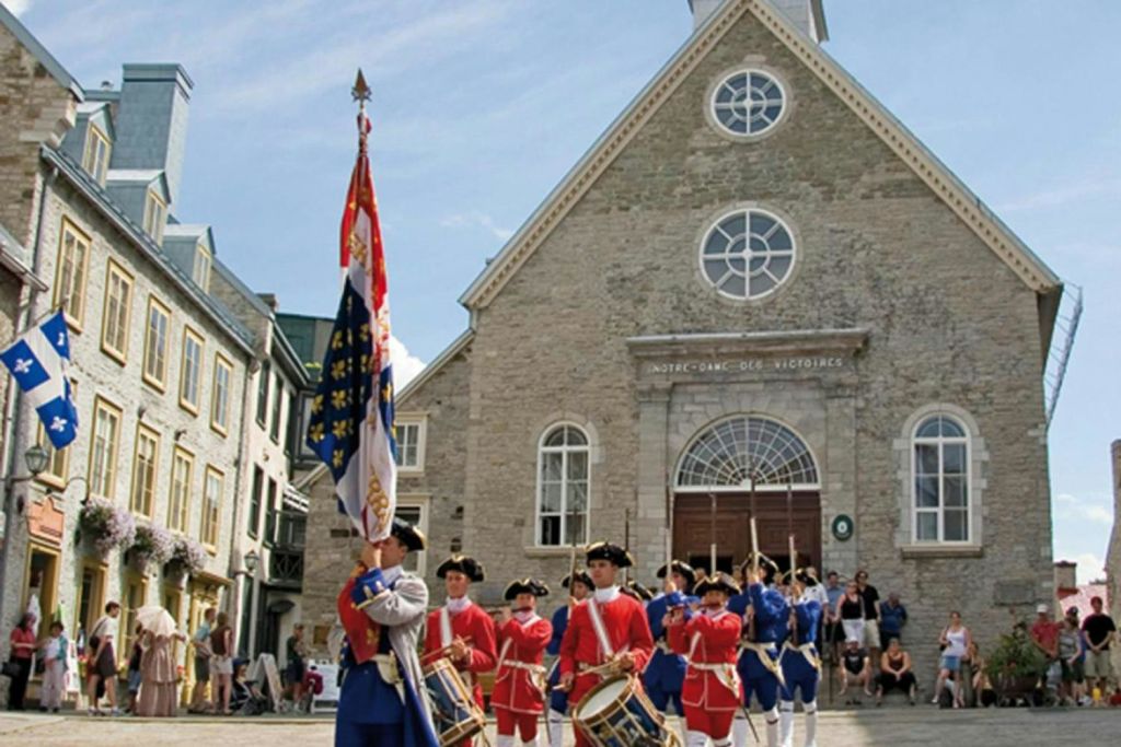 아치형 창문과 문이 있는 석조 교회 앞에서 붉은 유니폼을 입은 드러머와 깃발 소지자가 있는 역사적인 재연.