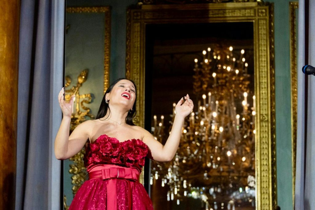 장미로 장식된 끈 없는 빨간 드레스를 입은 여성이 커다란 샹들리에를 배경으로 화려한 방에서 열정적으로 노래하고 있습니다.