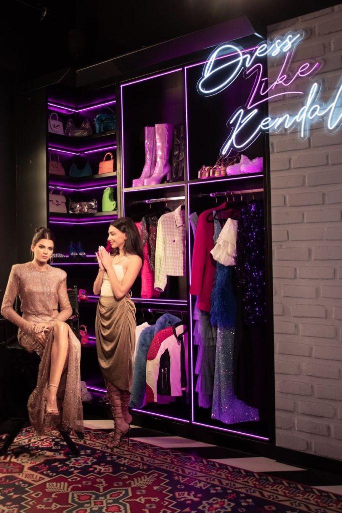 조명 옷장, 의류, 부츠, 가방, 네온사인이 있는 세련된 드레스룸에 두 명의 여성이 있습니다. 한 명은 앉아 있고, 다른 한 명은 박수를 치고 있습니다.