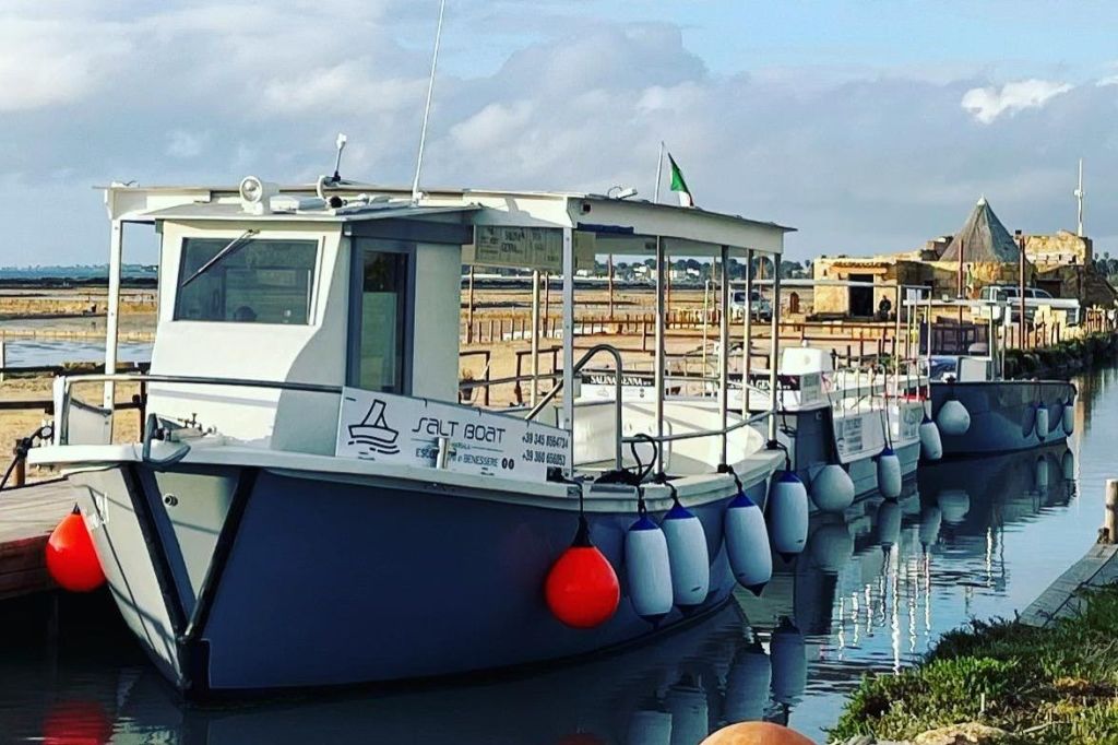 간판과 붉은 부표가 달린 흰색 보트가 잔잔한 물가에 정박해 있으며, 먼 해안선과 흐린 하늘을 배경으로 하고 있습니다.