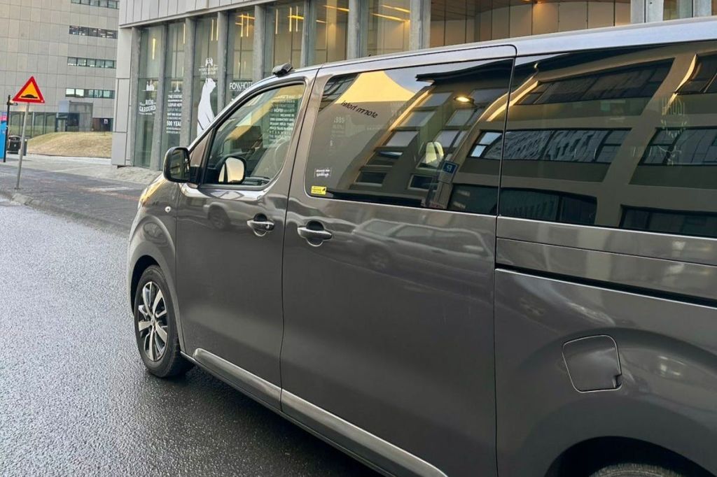 커다란 창문과 반사가 보이는 현대적인 건물 앞 젖은 거리에 회색 미니밴이 주차되어 있습니다.