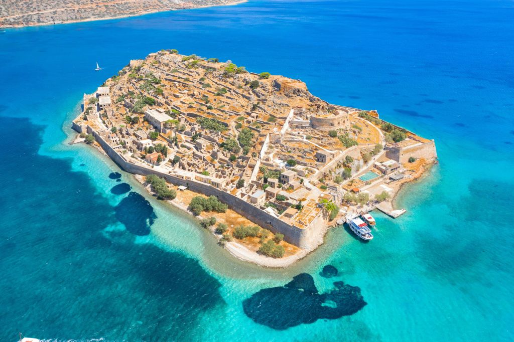 청록색 물로 둘러싸인 유적이 있는 요새화된 섬의 공중 전망. 해안에는 두 척의 보트가 정박해 있습니다.