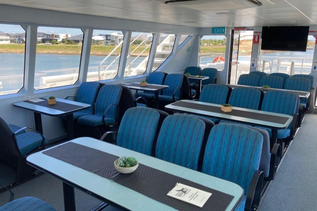 파란색 좌석, 테이블, 작은 화분, 출구 표지판이 있는 페리 내부. 물과 건물의 외부 전망.