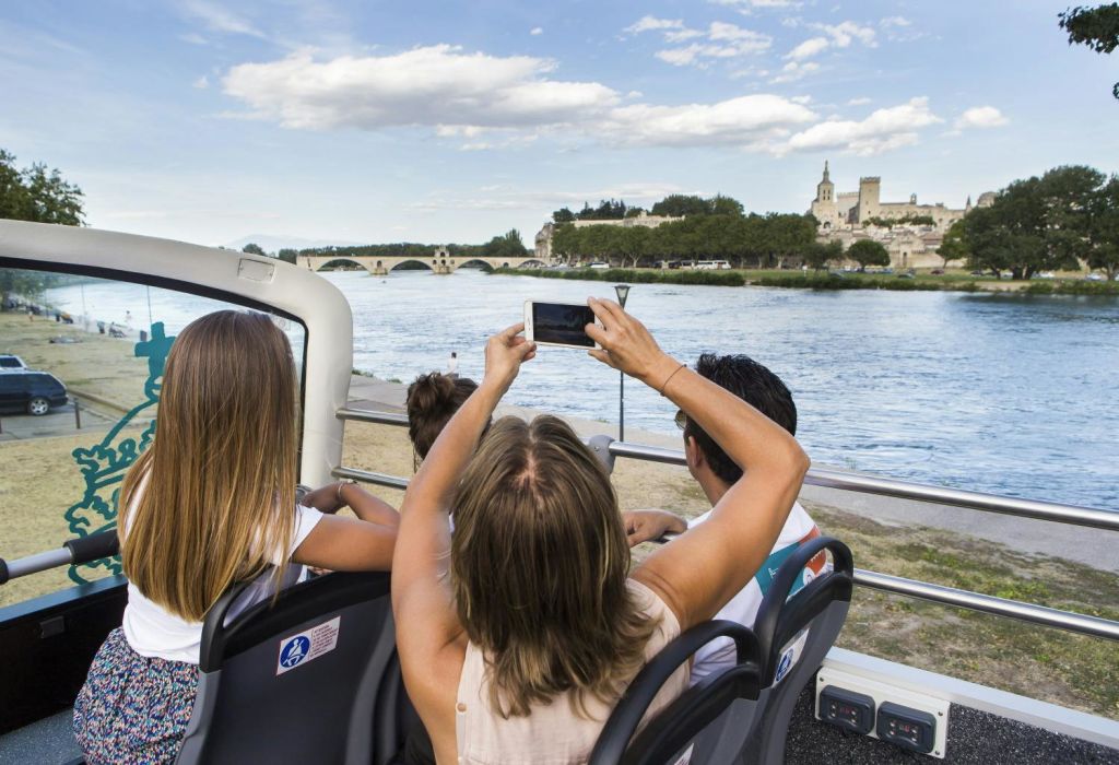 탁 트인 버스를 탄 세 사람이 구름이 낀 푸른 하늘 아래 역사적인 다리와 강을 배경으로 사진을 찍고 있습니다.