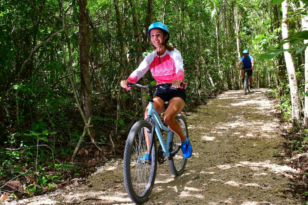 울창하고 푸른 숲을 가로지르는 흙길에서 자전거를 타고 헬멧을 쓰고 있는 두 사람. 앞에 있는 사람이 미소를 짓고 있습니다.