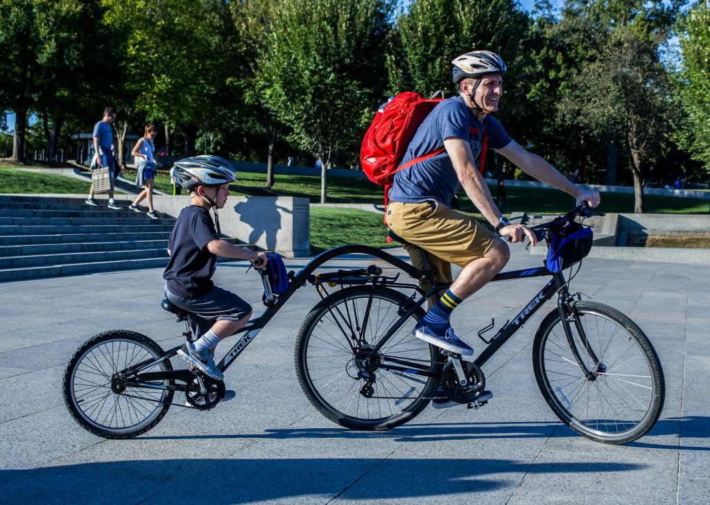 헬멧과 빨간색 배낭을 착용한 남성이 어린이 트레일러가 부착된 자전거를 타고 있습니다. 나무와 콘크리트 길이 배경입니다.
