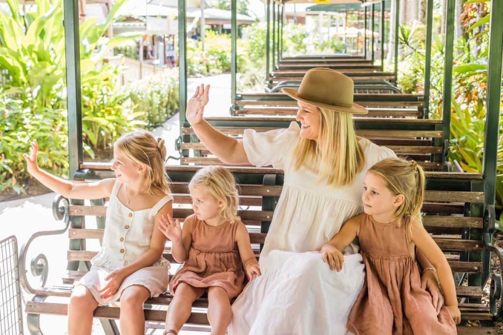 햇살이 내리쬐는 울창한 환경에서 녹색 야외 트램에 앉아 한 여성과 세 명의 소녀가 손을 흔들고 있습니다.