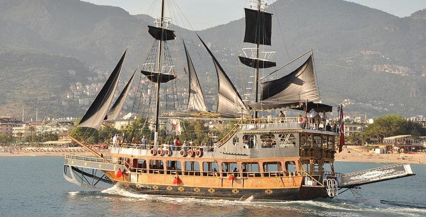 산과 건물로 둘러싸인 물 위에 검은 돛이 달린 목조 범선입니다. 사람들이 덱에 표시됩니다.