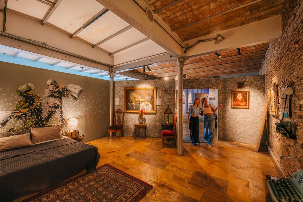 두 사람이 침대, 의자, 벽면 예술 작품, 노출된 벽돌 천장이 있는 예술 장식의 방을 거닐고 있습니다.