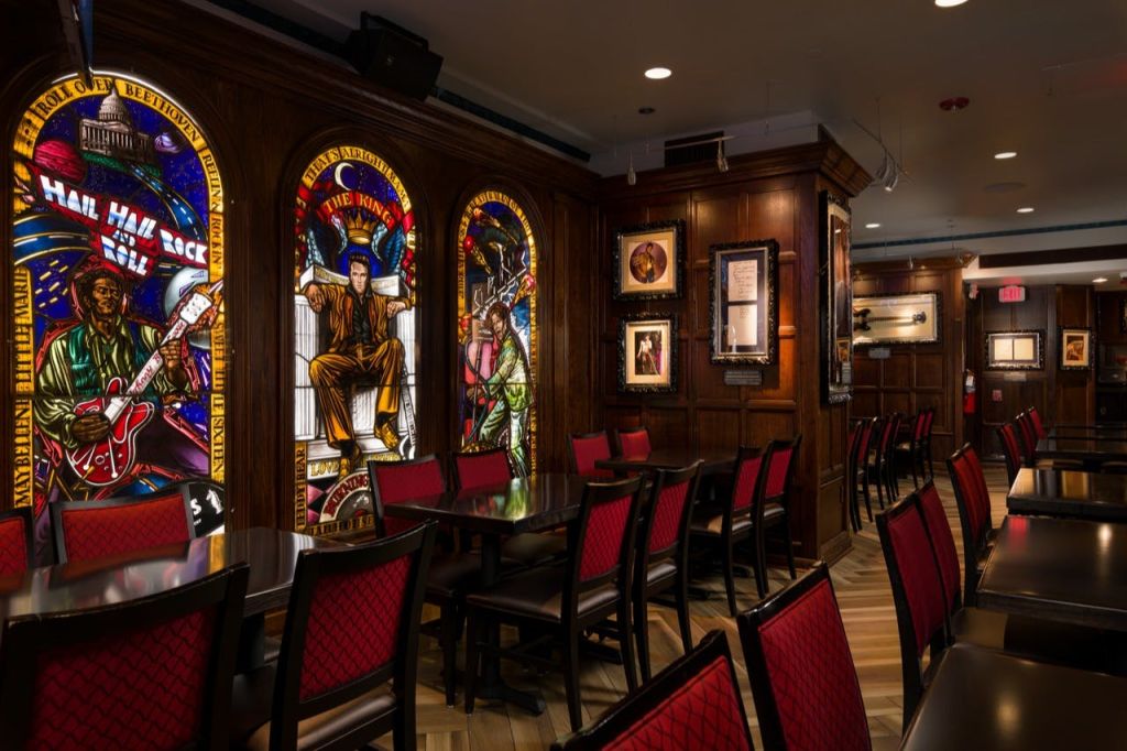 뮤지션을 묘사한 스테인드글라스 아트가 놓인 방, 벽에 액자 그림, 빨간색 실내 장식이 놓인 의자가 놓인 테이블.