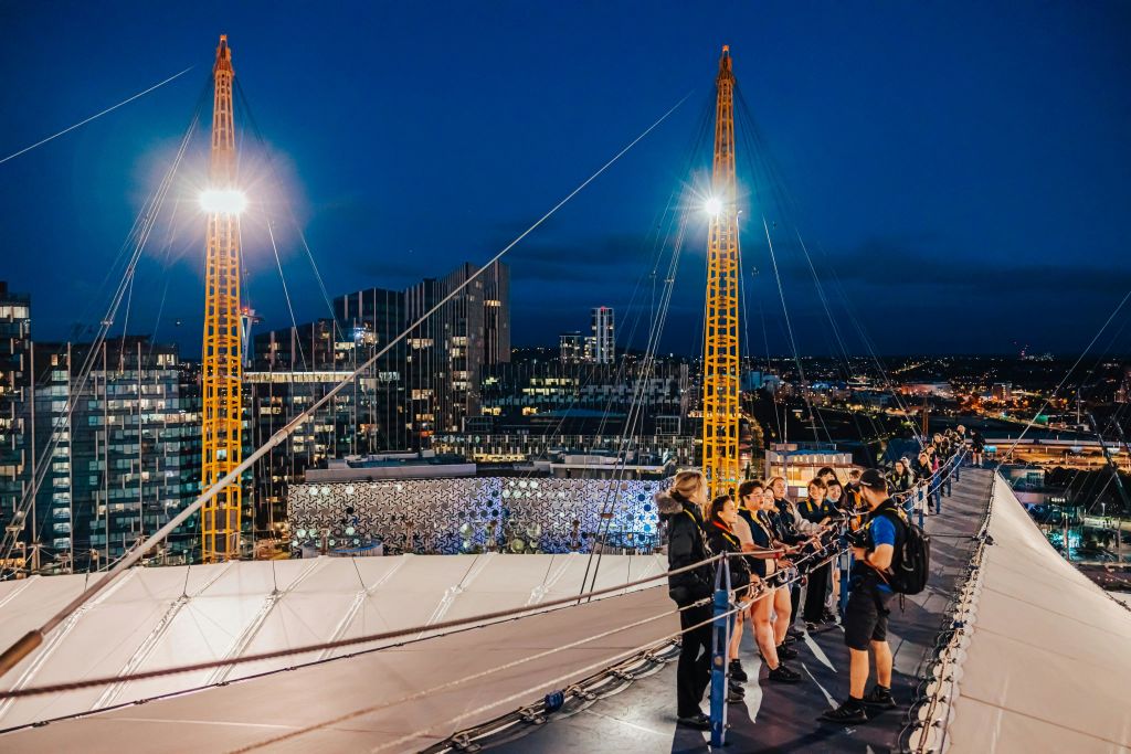 한 무리의 사람들이 건물과 조명 시설이 있는 도시 풍경을 내려다보며 밤에는 매달려 있는 통로에 서 있습니다.