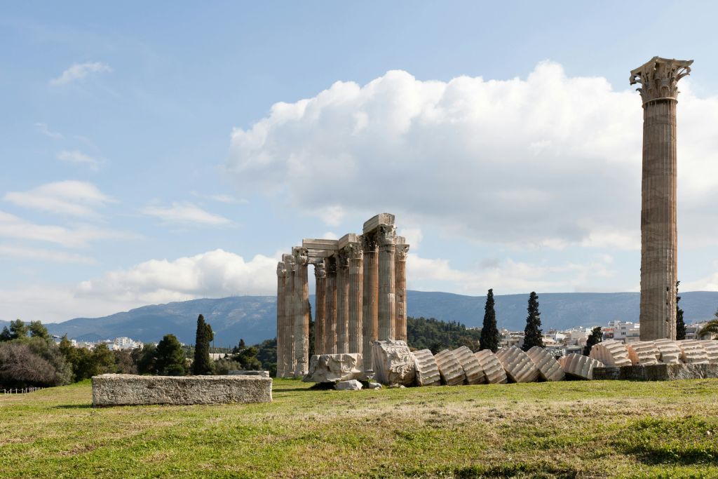 부분적으로 흐린 하늘 아래 산악 지대에 세워진 기둥과 떨어진 조각이 있는 고대 사원 유적.