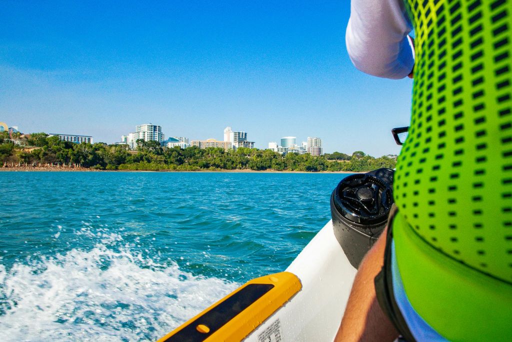 맑고 푸른 하늘 아래 건물의 도시 풍경 근처에서 물에 녹색 조끼를 입은 보트를 타고 있는 사람.