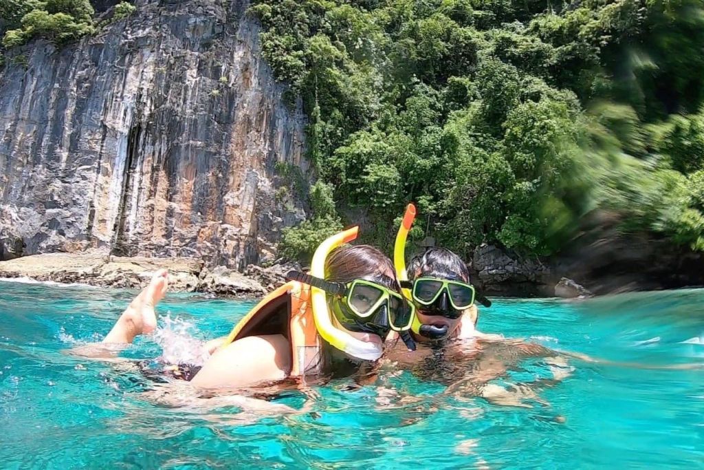 푸른 초목이 우거진 바위 절벽 근처의 청록색 맑은 물에서 스노클링을 즐기는 두 사람.