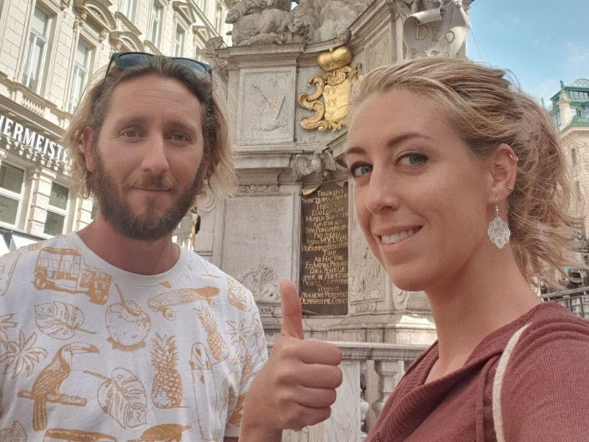 정교한 조각과 명판이 있는 기념비 앞에서 한 남녀가 셀카를 찍기 위해 포즈를 취하고 있습니다. 남자가 엄지손가락을 치켜세운다.