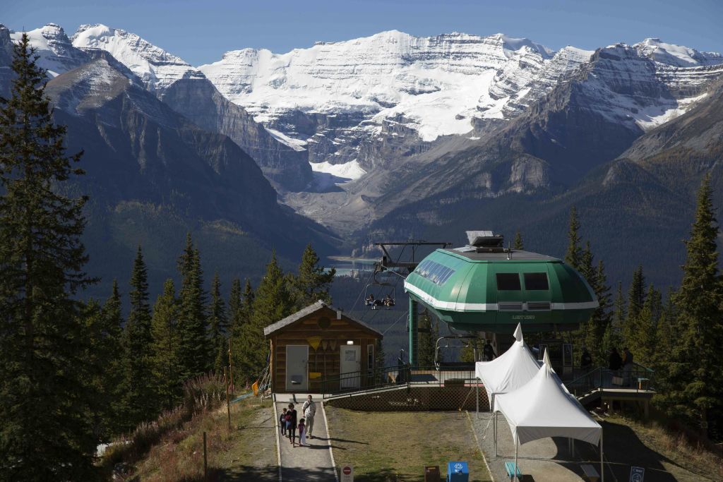 사람들은 눈 덮인 봉우리와 숲이 우거진 계곡을 배경으로 산의 녹색 곤돌라 정류장을 향해 걸어갑니다.