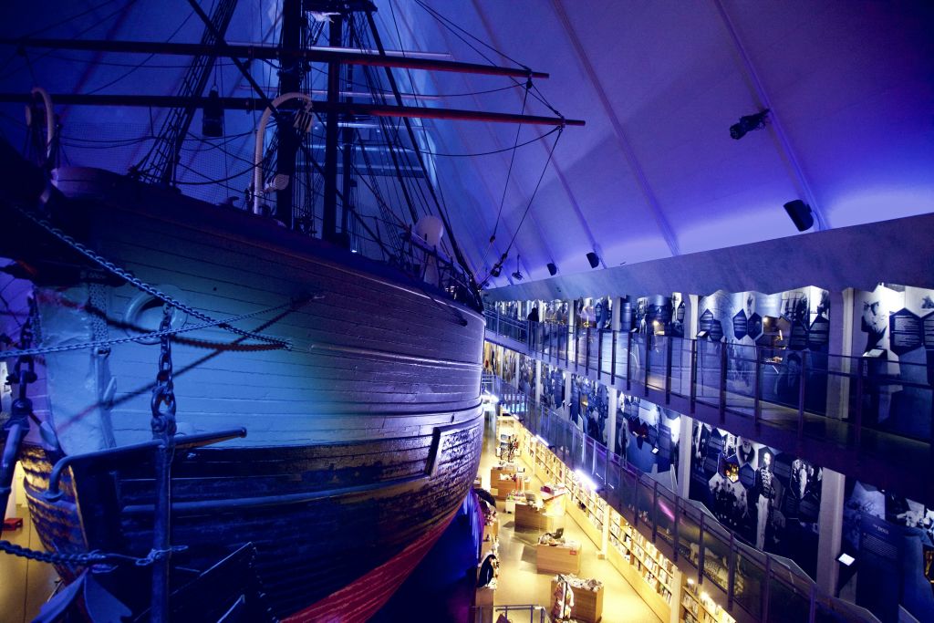 역사적인 전시물과 방문객이 있는 2층 갤러리 옆의 파란색 조명 아래 실내에 대형 목조선이 전시되어 있습니다.