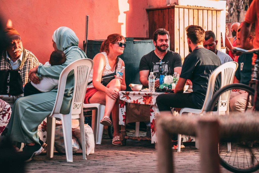 4명이 야외 작은 테이블 주위에 하얀 의자에 앉아 대화에 참여하고 다양한 물건으로 둘러싸여 있습니다.