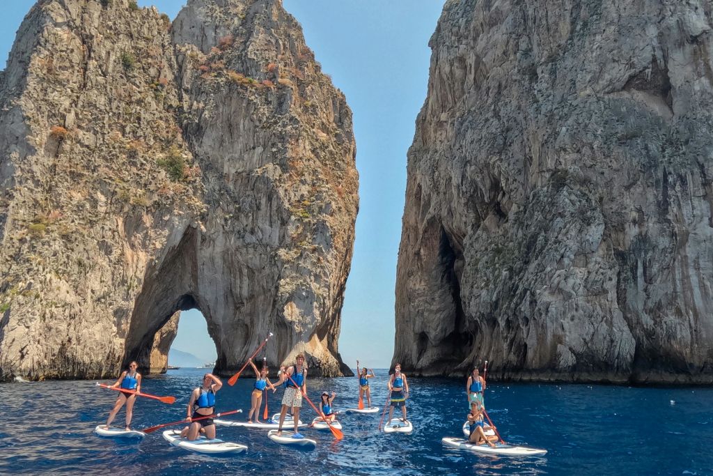 화창한 날 자연의 아치가 있는 커다란 암석 지층 근처에서 푸른 물 위에서 패들보딩을 즐기는 사람들.
