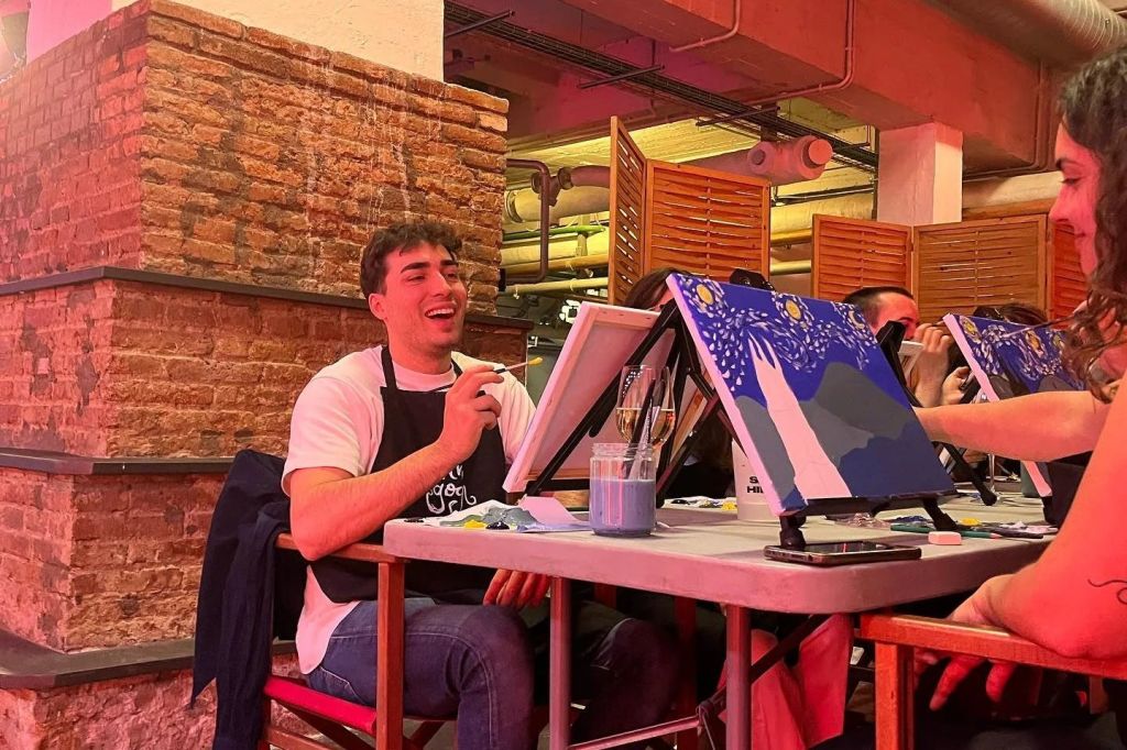 흰색 셔츠와 앞치마를 입은 사람이 인더스트리얼 스타일의 공간 안에 있는 이젤에서 그림을 그리며 미소를 짓고 있습니다.