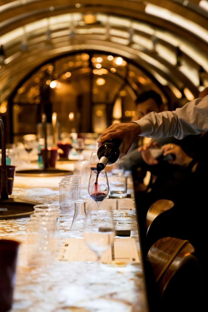 한 사람이 아늑하고 아치형으로 꾸며진 실내 환경에 놓인 길고 밝은 테이블에 레드 와인을 붓고 있습니다.