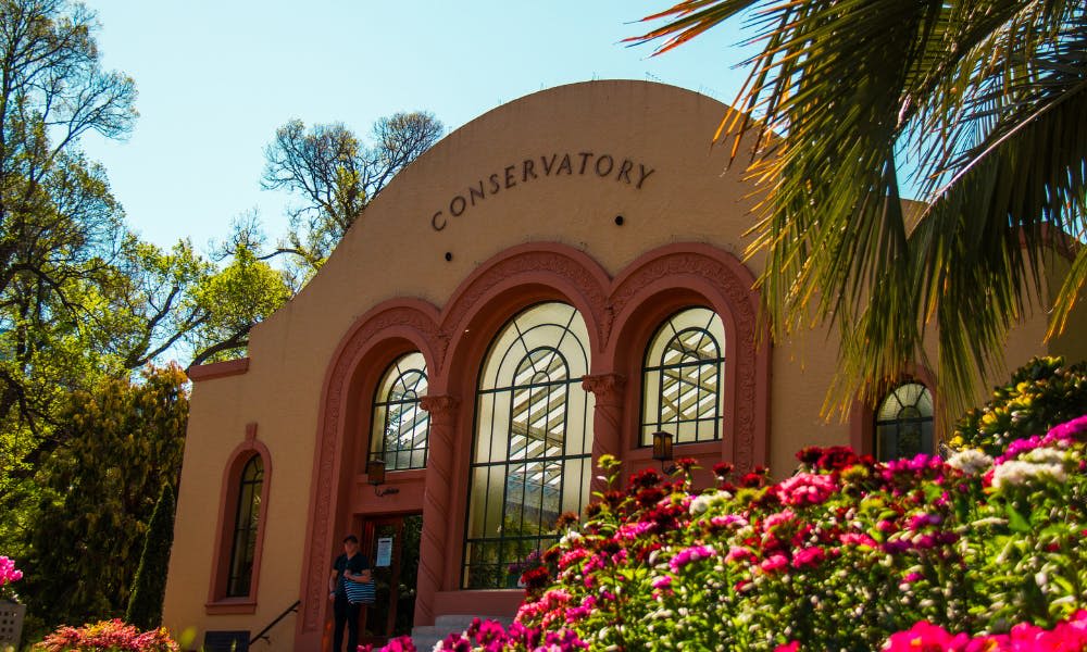 "온실" 이라고 표시된 베이지색과 분홍색 아치형 건물로, 커다란 아치형 창문이 있으며 꽃과 녹음으로 둘러싸여 있습니다.