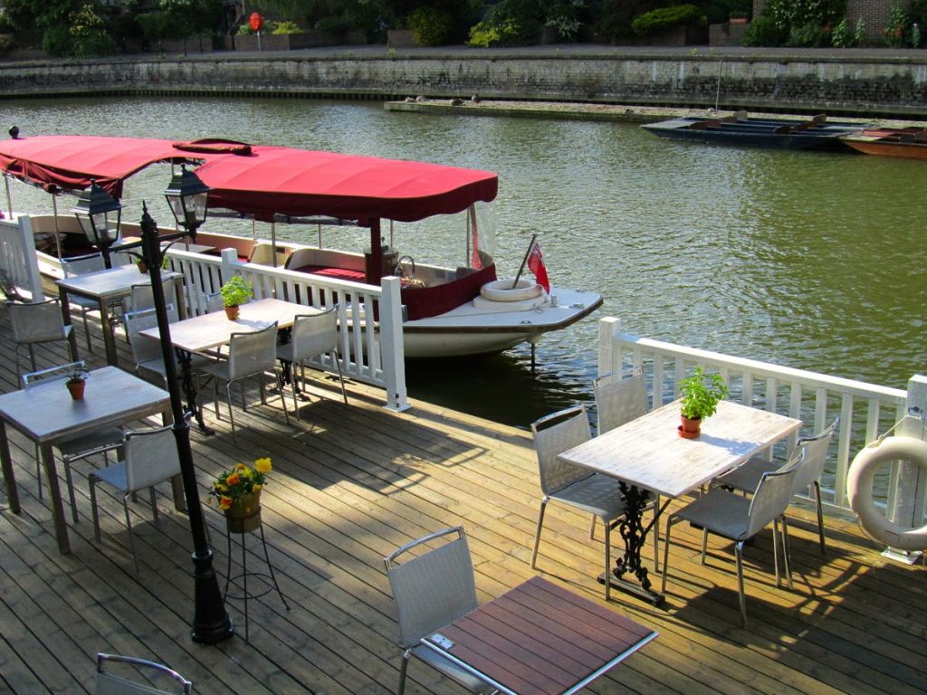 보트가 있는 운하가 내려다보이는 의자와 테이블이 있는 야외 나무 데크 (빨간색 캐노피로 그늘진 데크 포함).