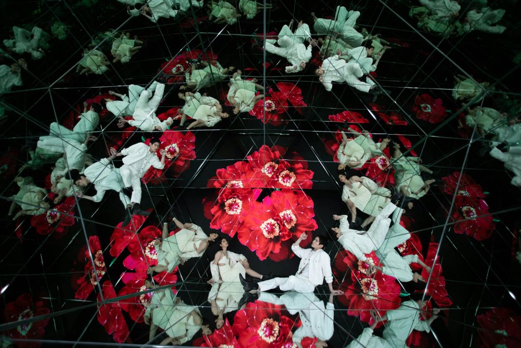 흰색 의상을 입은 두 사람이 만화경 패턴의 커다란 붉은 꽃을 반사하는 거울로 둘러싸여 있습니다.