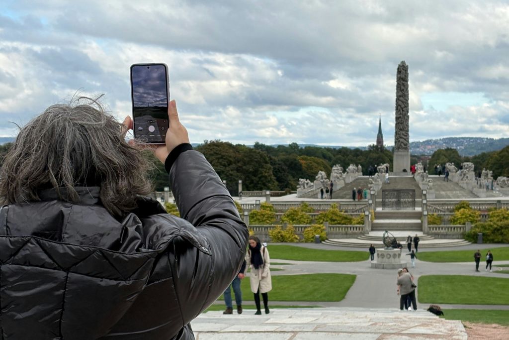 검은 코트를 입은 사람이 동상, 녹지, 흐린 하늘 아래 오벨리스크가 있는 공원을 촬영하고 있습니다.