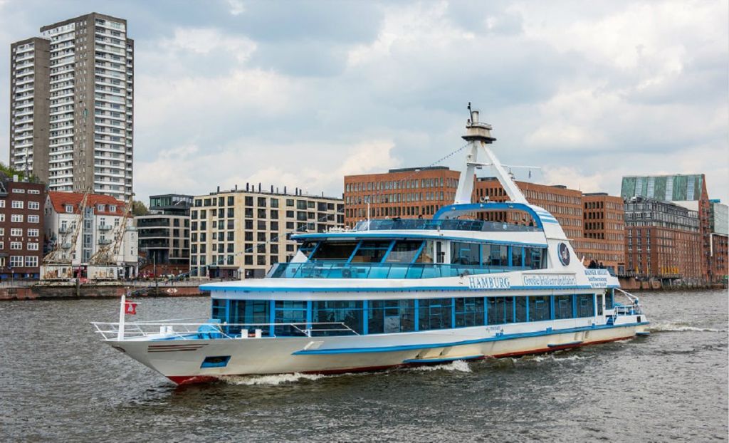 '함부르크' 라는 이름의 파란색과 흰색 여객선이 현대적인 건물을 배경으로 강을 거닐고 있습니다.
