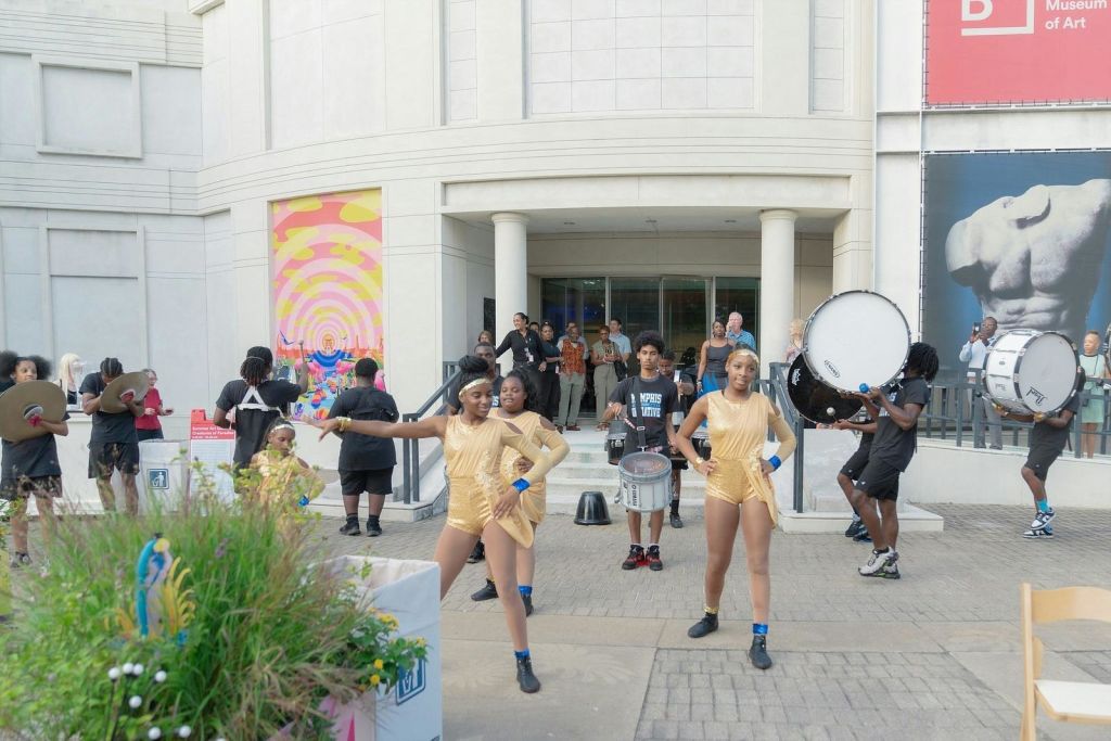 금색 의상을 입은 댄스 그룹이 '미술관' 안내판이 있는 건물 밖에서 드러머와 함께 공연합니다. 사람들은 배경을 보고 있습니다.