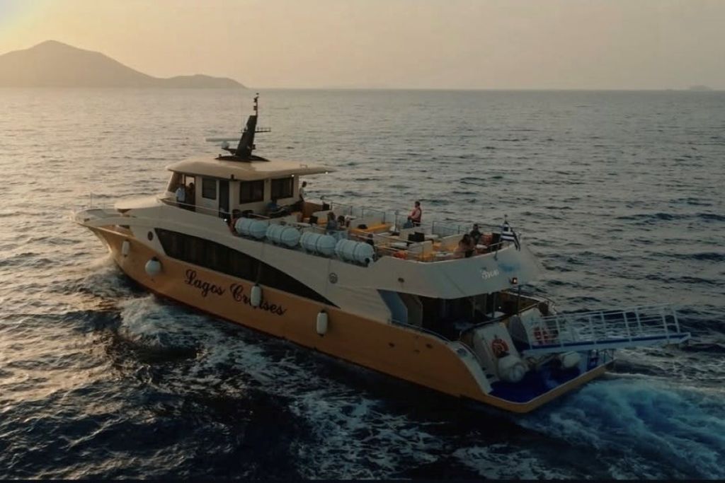 '라고스 크루즈 (Lagos Cruises)' 라는 이름의 크루즈 보트는 먼 해안선 근처에서 일몰이 지는 동안 갑판에 있는 사람들과 함께 잔잔한 물 위를 항해합니다.