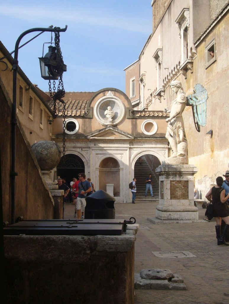 돌상, 걷는 사람들, 아치형 출입구, 흉상이 있는 건물로 올라가는 계단이 있는 안뜰.