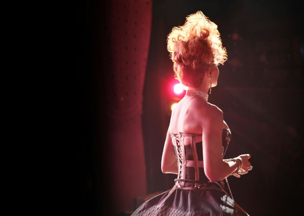 곱슬머리를 한 여성이 무대 위에 서서 코르셋과 초커를 착용하고 극적인 조명으로 실루엣을 강조합니다.