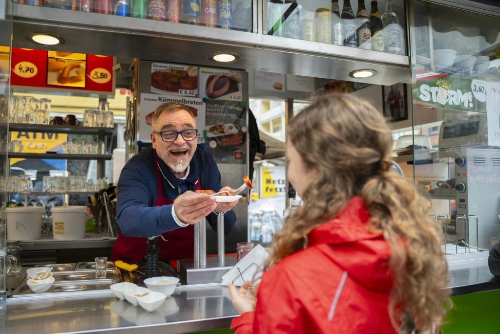 음식 가판대에서 미소를 짓는 상인이 빨간 재킷을 입은 고객에게 요리를 제공합니다. 위에 다양한 메뉴 항목이 표시됩니다.