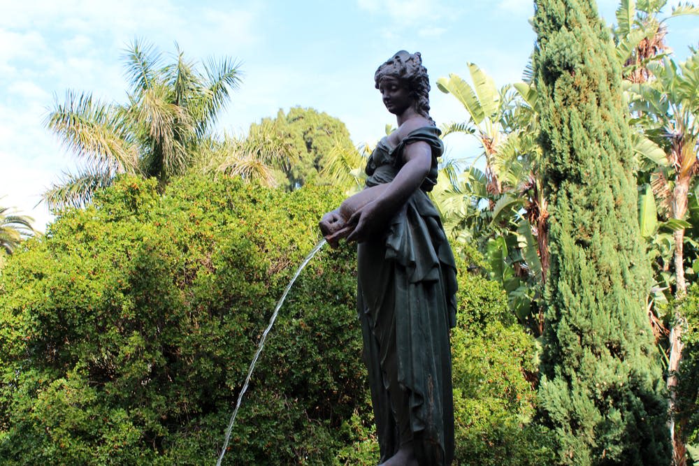 푸른 하늘 아래 푸른 녹음과 다양한 나무들로 둘러싸인 주전자에서 물을 붓는 여성의 동상.