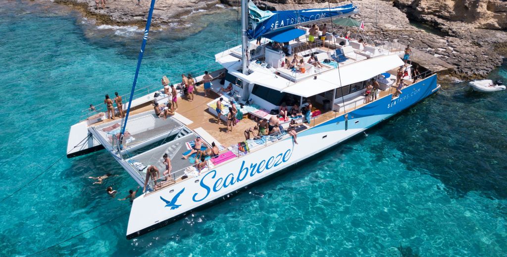 바위가 많은 해안가 근처의 맑고 청록색 바다에서 일광욕을 즐기고 휴식을 취하며 사람들과 어울리는 "바닷바람" 이라는 이름의 대형 쌍동선.