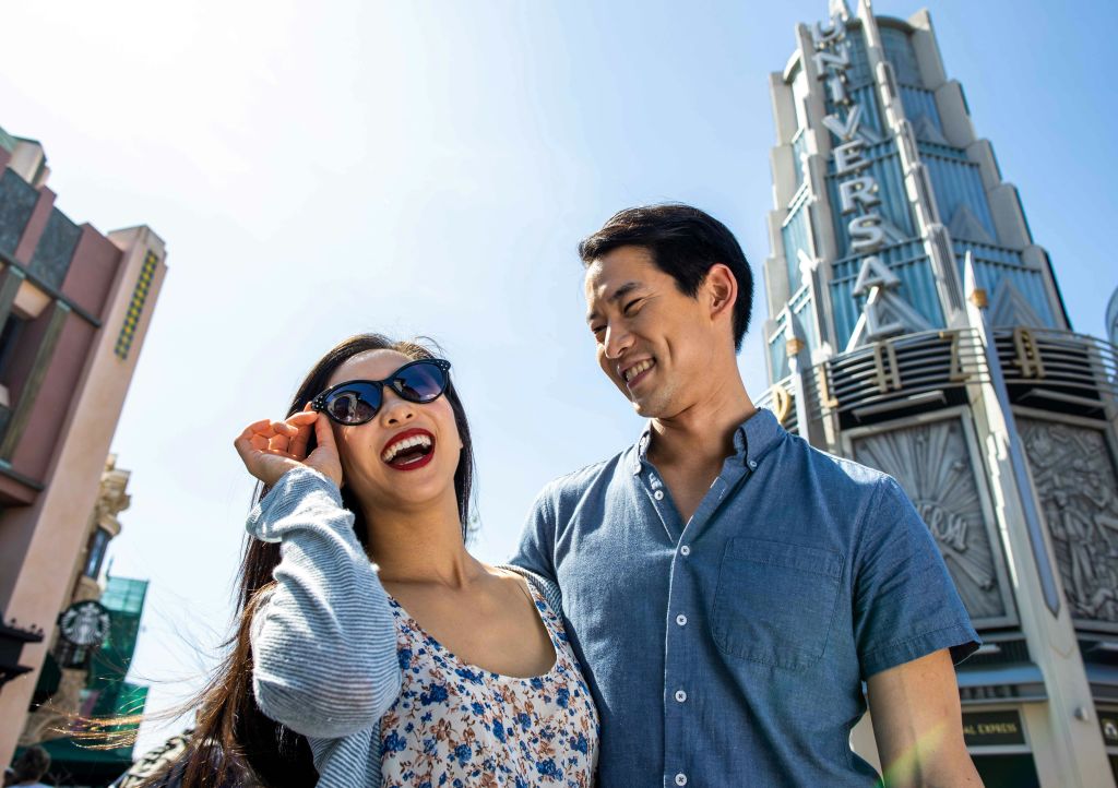 맑고 화창한 날, '유니버설' 이라고 적힌 우뚝 솟은 표지판 앞에 미소를 짓는 선글라스를 쓴 부부가 서 있습니다.